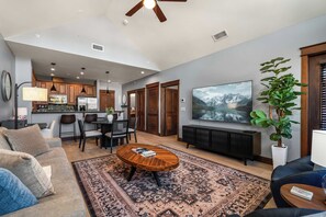 A modern living room with a large TV, a coffee table, and a sofa. There is a kitchen area in the background with wooden cabinets and a dining table. - Cozy Riverstone Escape | Walkable CDA Location (Coeur d'Alene)