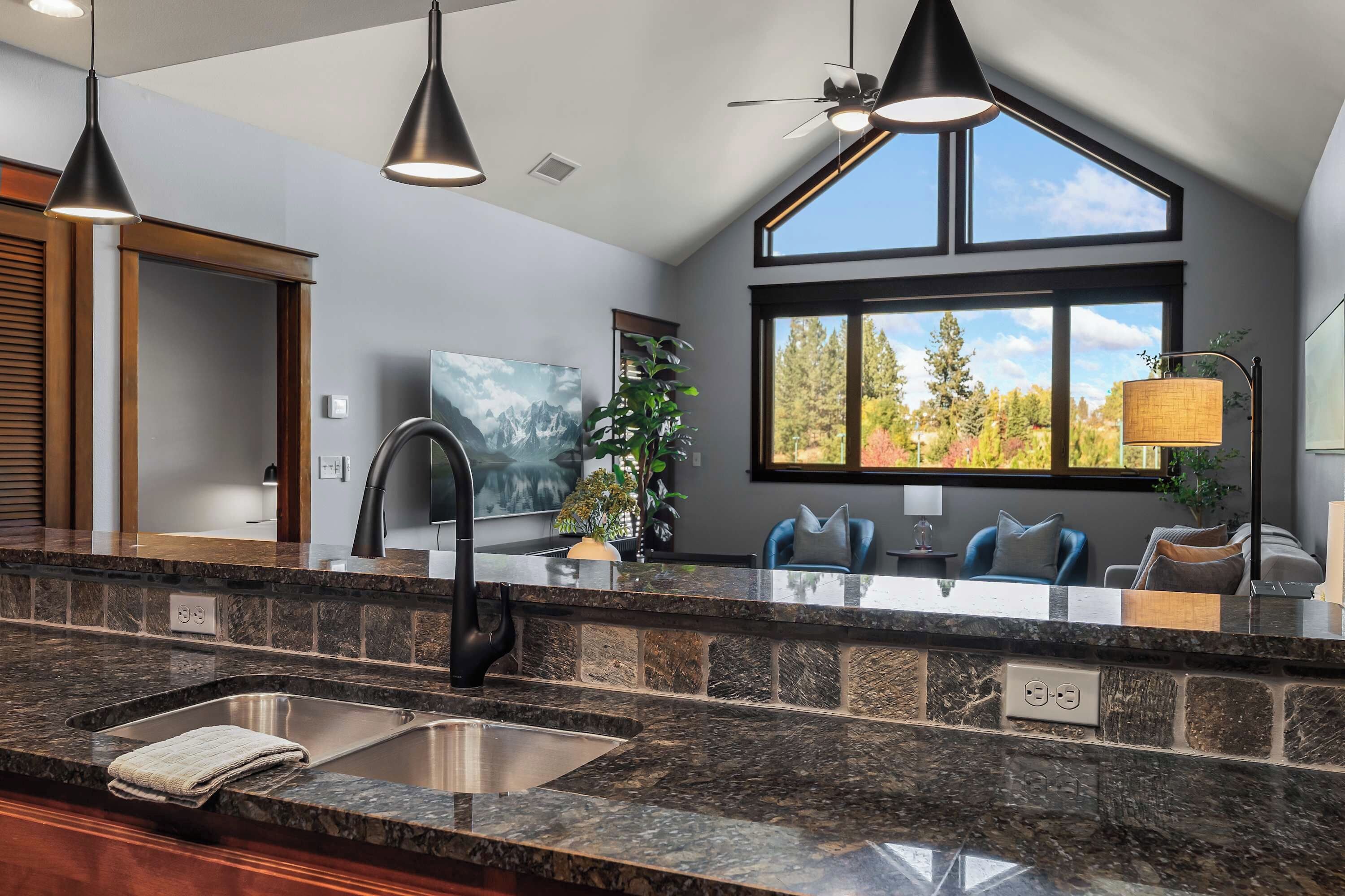 A modern kitchen with granite countertops, a double sink, a built-in oven, and a large window with a view of trees.