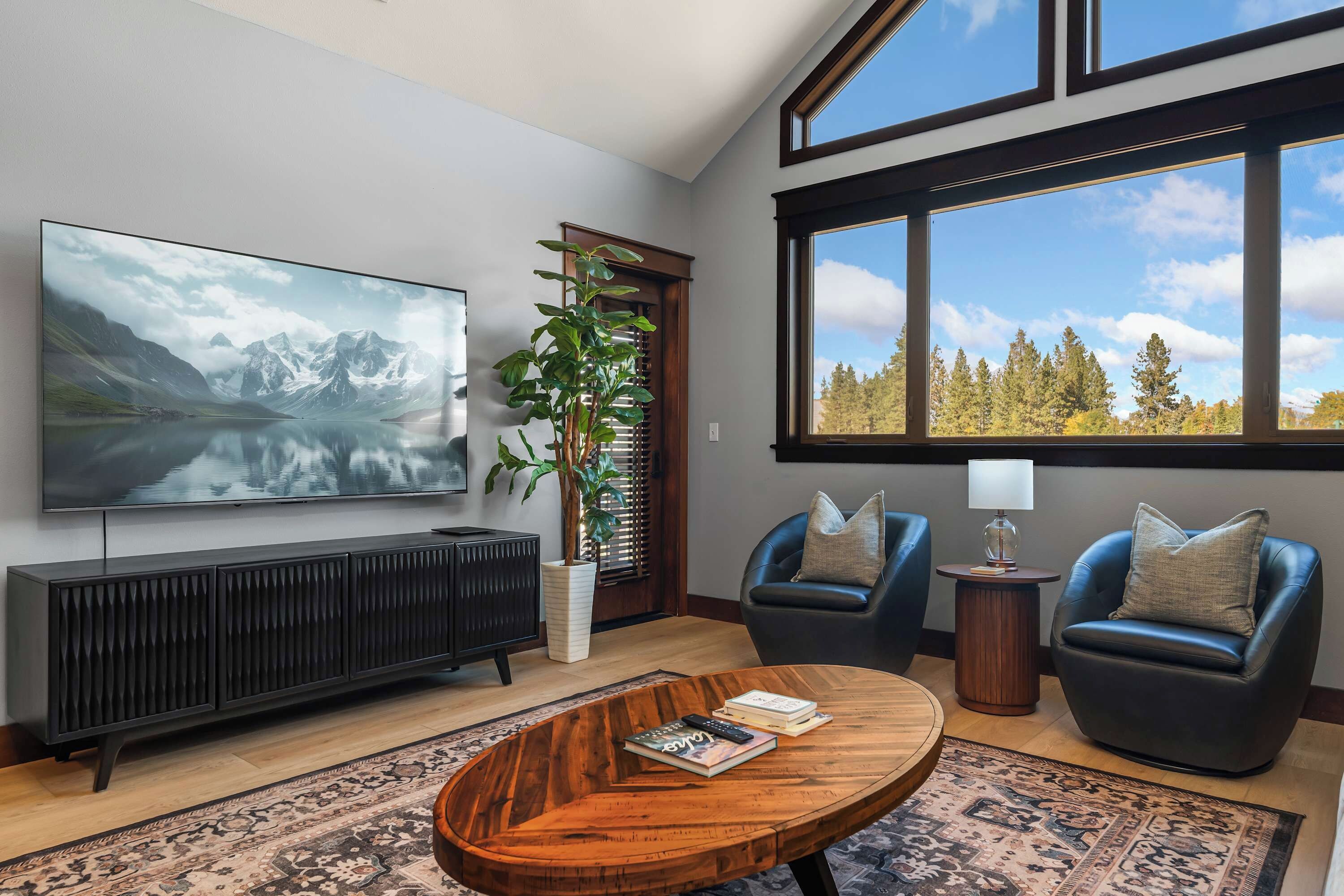 A modern living room with a large TV, two leather chairs, a coffee table, and a view of a lake and forest through large windows.