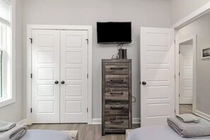 A bedroom with two white doors, a wooden dresser, a flat-screen TV, and a bed with a gray blanket. - Beach Retreat Near Pawleys Island w/ Open Living, Deck & Resort Amenities (Pawleys Island)