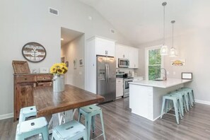A modern kitchen with white cabinets, stainless steel appliances, and a wooden dining table with light blue bar stools. - Beach Retreat Near Pawleys Island w/ Open Living, Deck & Resort Amenities (Pawleys Island)