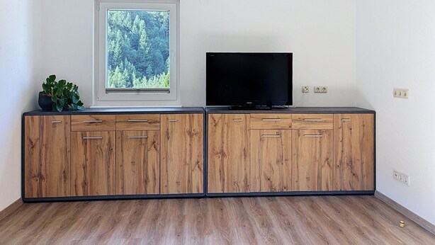 Ein Zimmer mit einem Holzkasten, einem Fernseher darauf, einem Fenster mit Blick auf einen Wald und einer Pflanze auf dem Kasten.