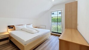 Ein Schlafzimmer mit einem Bett, einem Nachttisch, einem Holztisch und einem großen Fenster mit Blick auf Grünflächen.