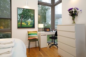 Ein Schlafzimmer mit einem Bett, einem Schreibtisch mit Computer, einem Sessel, einer Lampe und einem Fenster mit Blick auf Bäume.