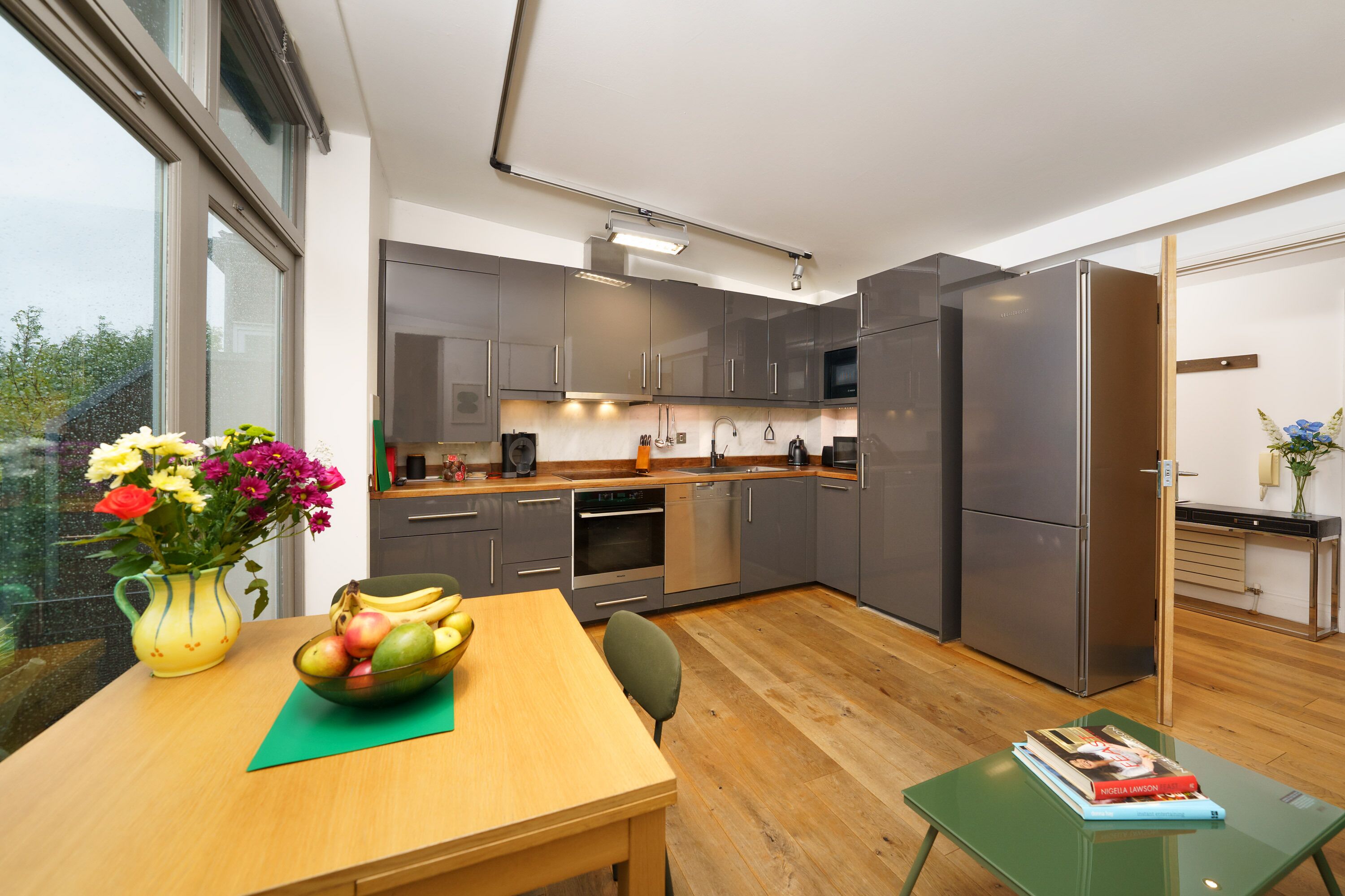 Cocina moderna con armarios grises, electrodomésticos de acero inoxidable y una mesa de comedor de madera con un frutero y un florero.