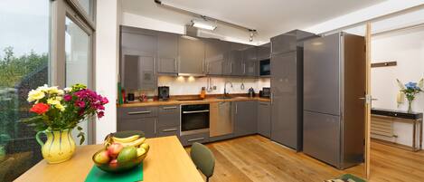 A modern kitchen with grey cabinets, stainless steel appliances, and a wooden dining table with a fruit bowl and a vase of flowers.