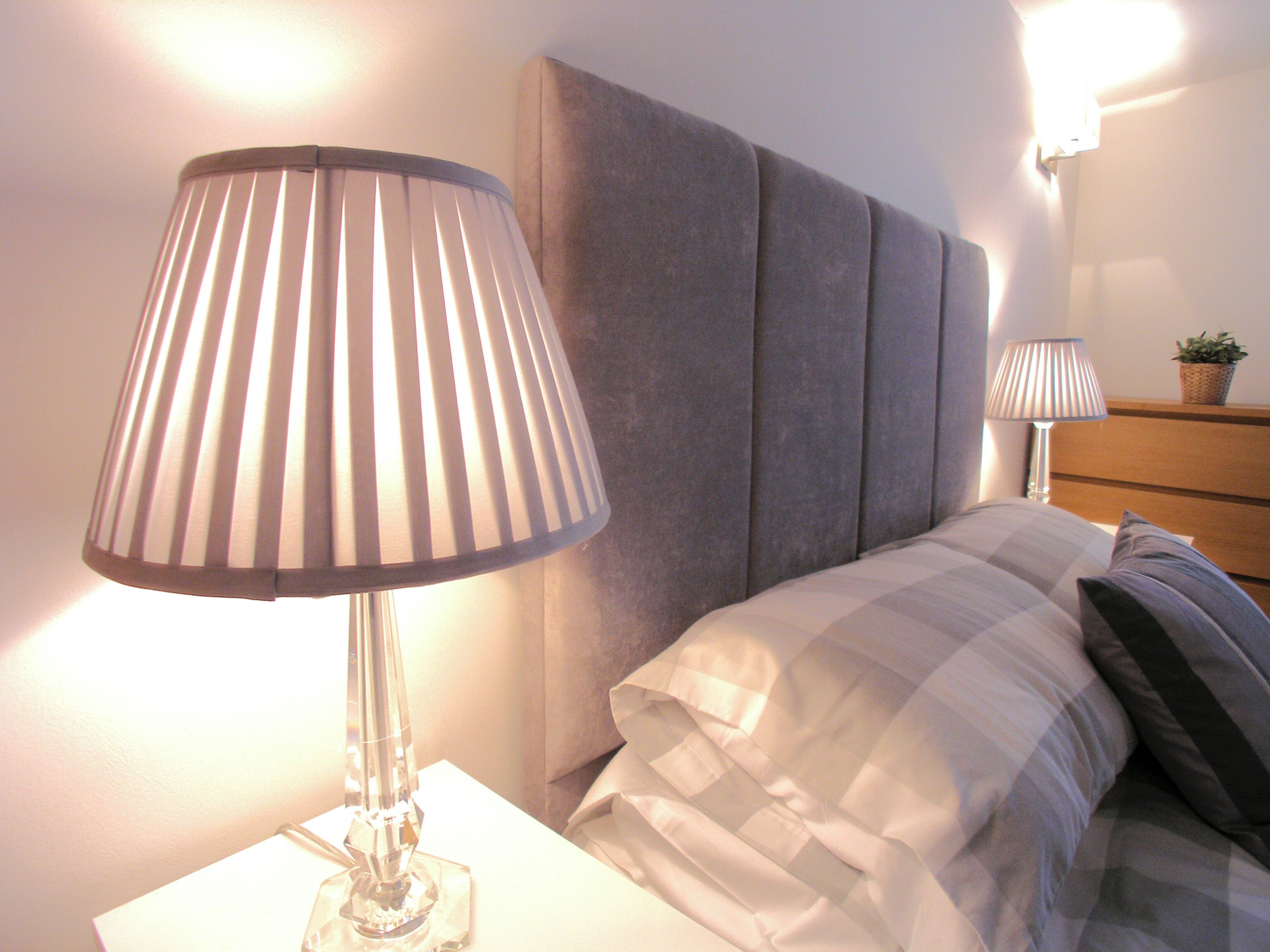 A neatly made bed with a grey headboard and white bedding, a bedside table with a lamp, and a small potted plant on the nightstand.