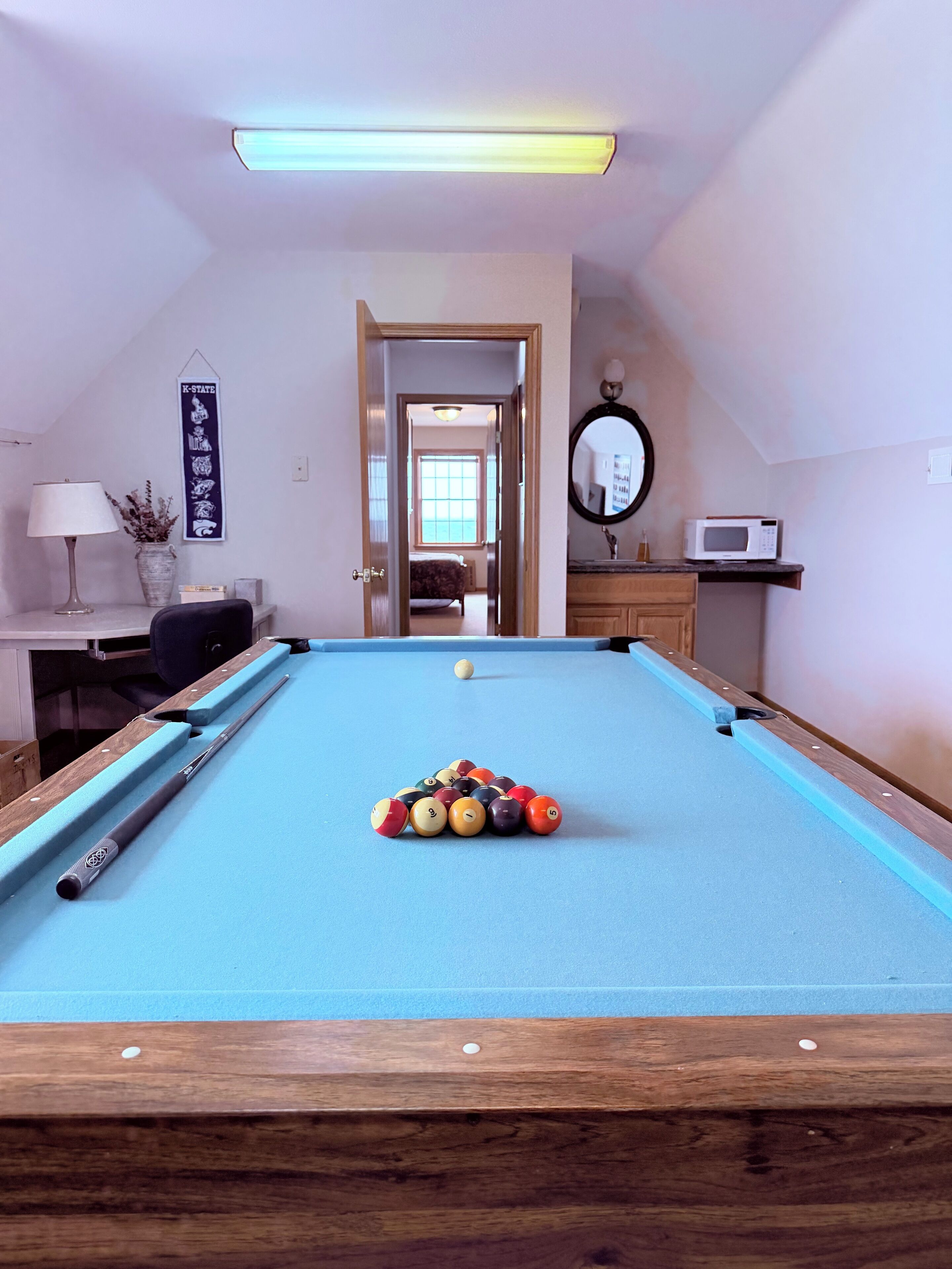 Una mesa de billar con bolas, una vara de billar, una lámpara, un florero con plantas, un espejo y un microondas en una habitación con una puerta que da a otro espacio.