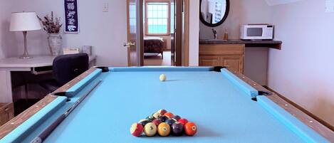 Una mesa de billar con bolas, una vara de billar, una lámpara, un florero con plantas, un espejo y un microondas en una habitación con una puerta que da a otro espacio.