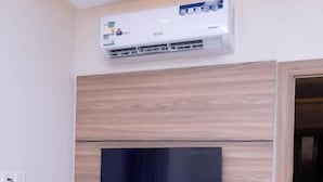 Una habitación con televisor de pantalla plana montado, paneles de madera en las paredes, unidad de aire acondicionado integrada y un pequeño escritorio con teléfono.