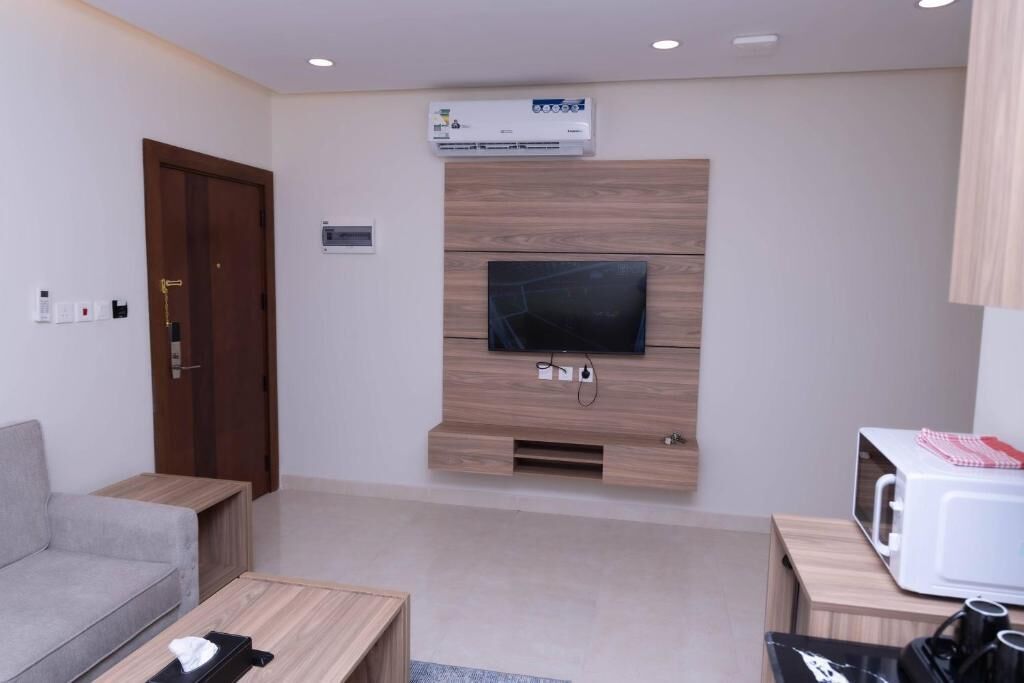 A modern living room with a flat-screen TV mounted on a wooden wall, a grey sofa, a wooden coffee table, and a microwave on a wooden cabinet.