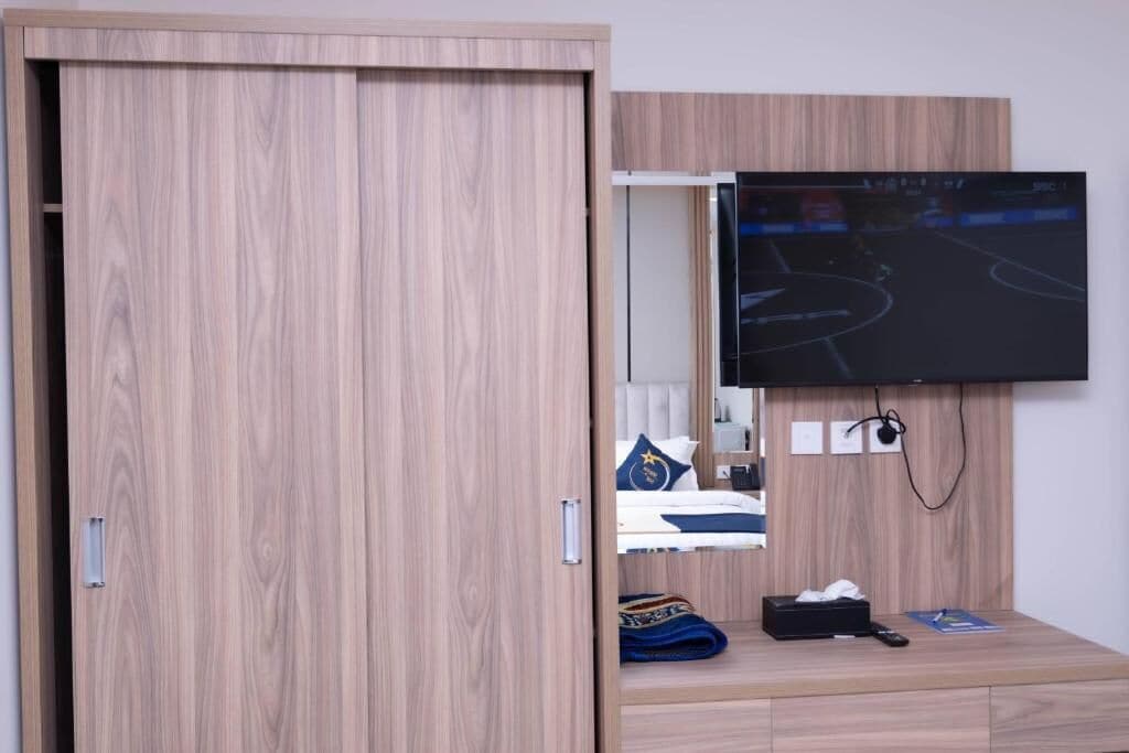 A room with a wooden wardrobe, a bed with a headboard, a television mounted on the wall, and a small table with items on it.