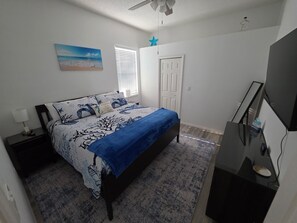 Ein Schlafzimmer mit Bett, Nachttisch, Schreibtisch, Stuhl, Fernseher, einem Fenster mit Jalousien und einer Tür.