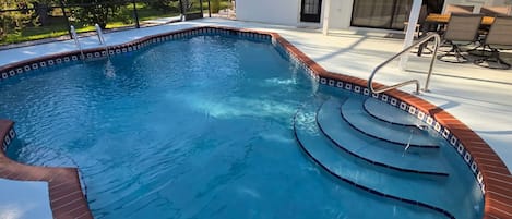 A swimming pool with a brick border, a covered area with chairs, and a door leading to a covered patio.