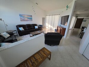 Ein geräumiges Wohnzimmer mit einer Couch, einem Couchtisch und einer Holzbank. Ein Gemälde hängt an der Wand und durch ein Fenster bietet sich ein Blick ins Freie.