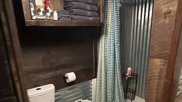 Un baño pequeño con inodoro, estante con toallas dobladas, cortina de ducha y un taburete.