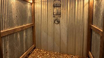 A wooden outdoor shower area with a corrugated metal wall, a stone floor, and a metal rack with bottles.