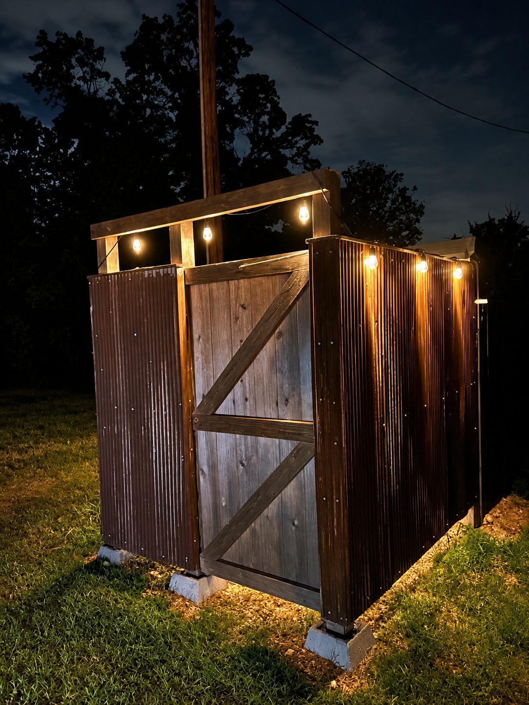 A wooden structure with a metal gate, illuminated by string lights, set on a grassy area.