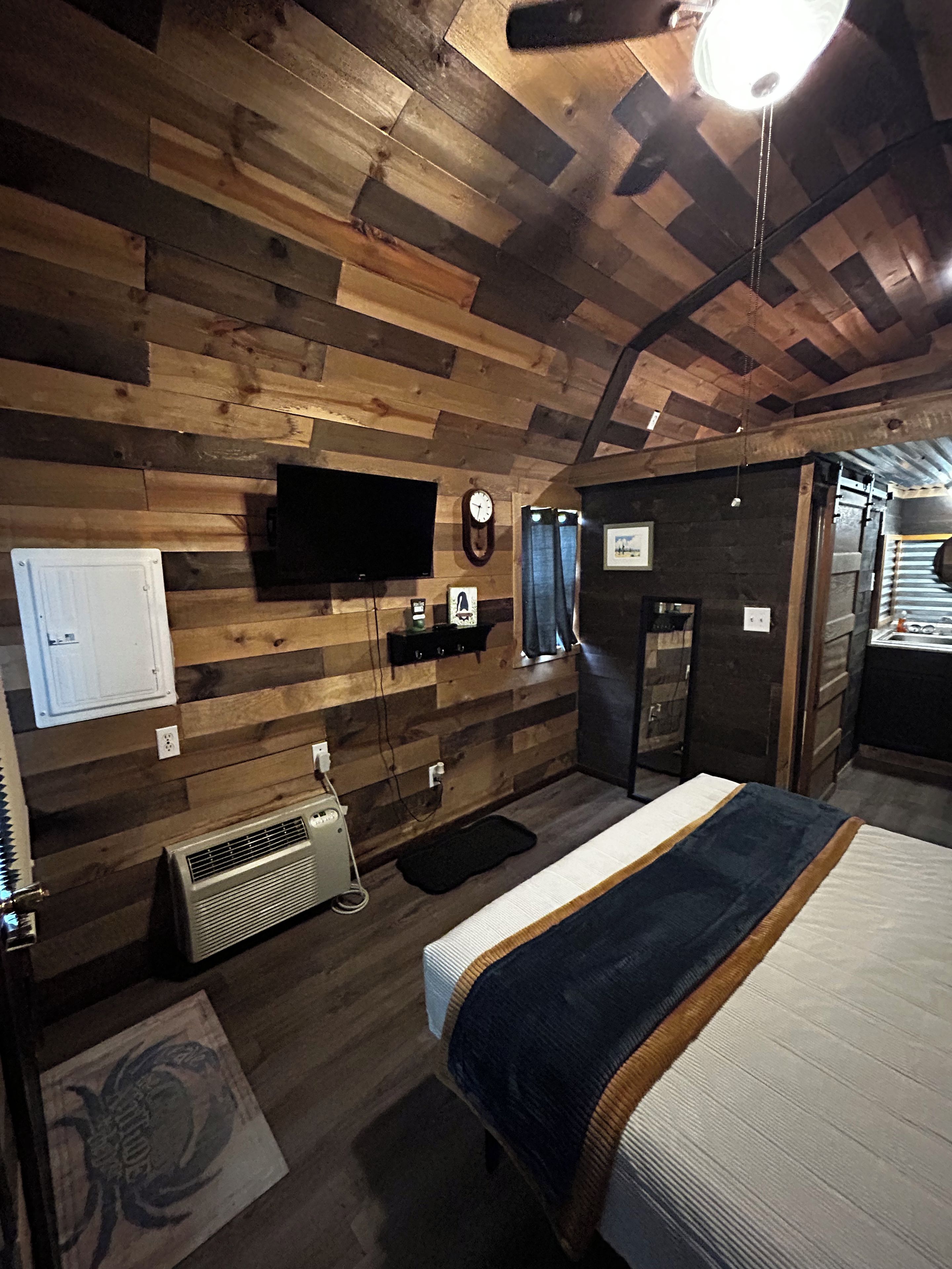 Una habitación con techo de madera, cama con colcha azul y blanca, televisor montado, un reloj y un calentador.
