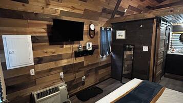 Una habitación con techo de madera, cama con colcha azul y blanca, televisor montado, un reloj y un calentador.