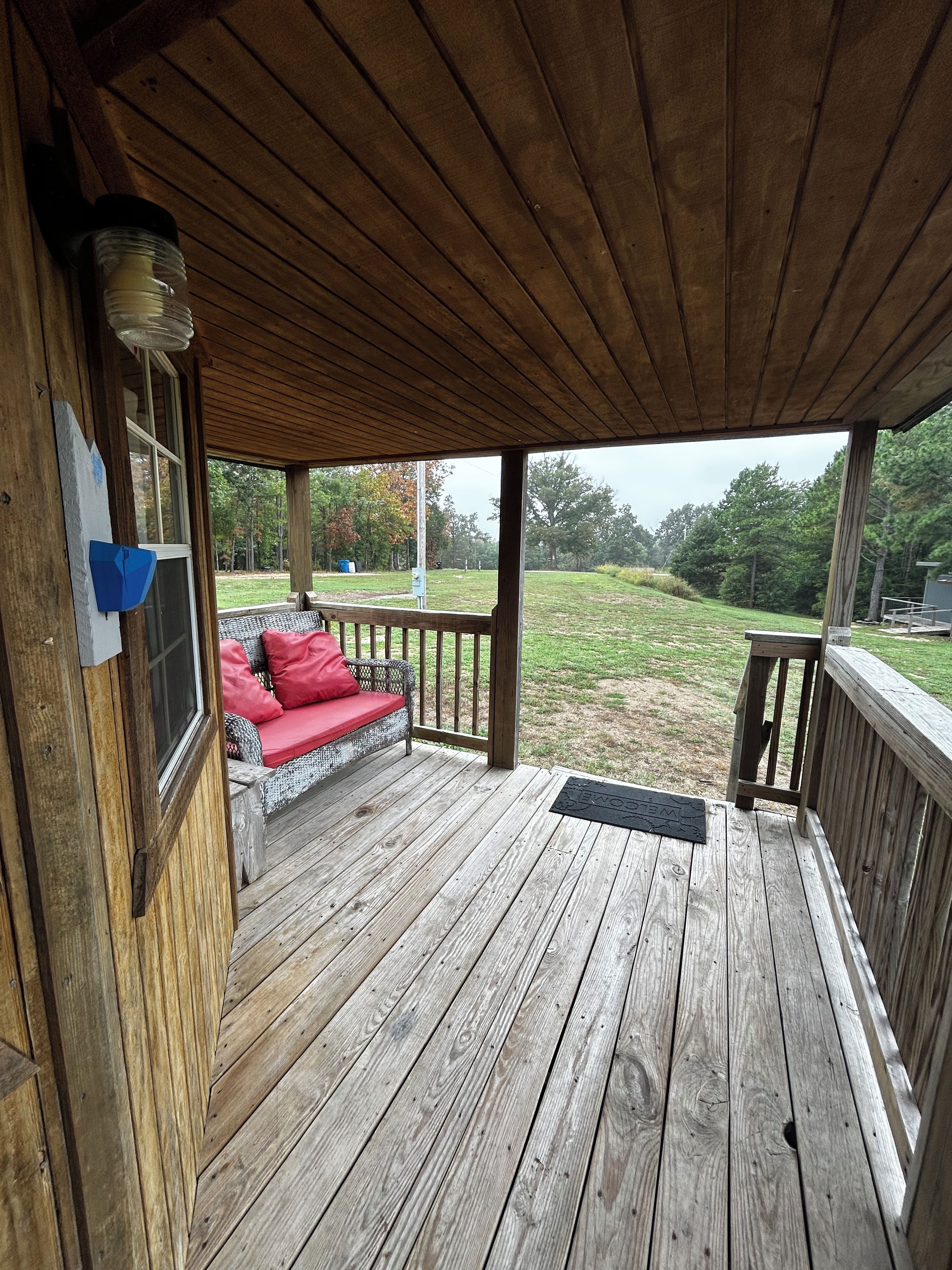 A wooden deck with a red cushioned bench, a doormat, and a view of a grassy area with trees.