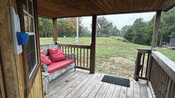A wooden deck with a red cushioned bench, a doormat, and a view of a grassy area with trees.