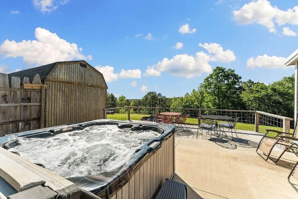 A hot tub with a wooden deck, a table, and chairs, surrounded by a fence and trees.