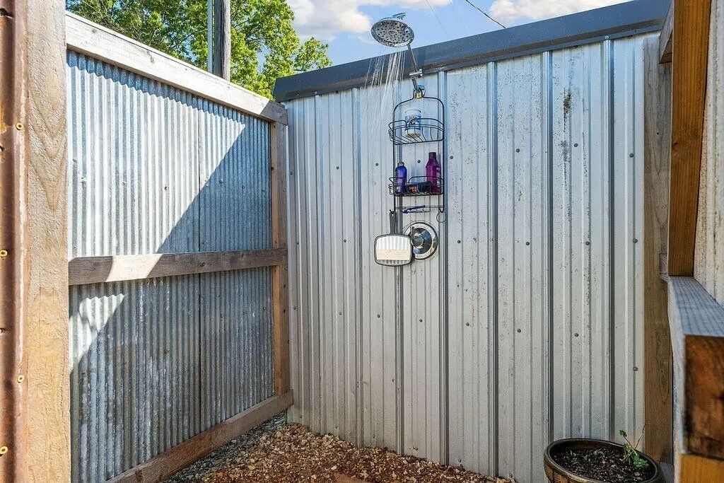 A metal-walled outdoor shower area with a hose, a water container, and a shelf with bottles.