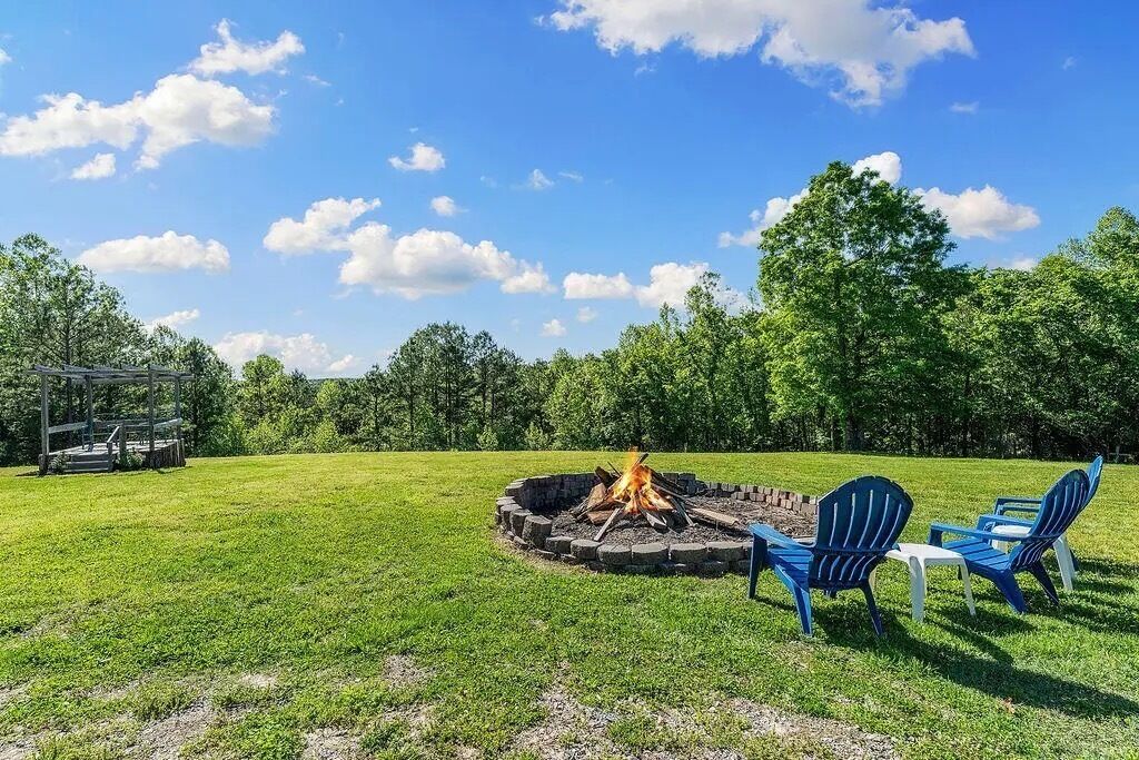 A circular fire pit with chairs and a table, surrounded by a grassy field and trees.