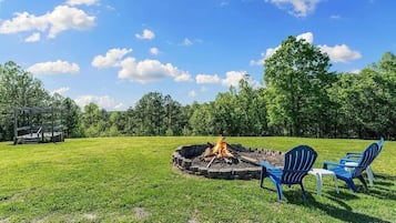 A circular fire pit with chairs and a table, surrounded by a grassy field and trees.