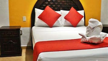 A hotel room with a double bed, red pillows, a red throw blanket, and a towel folded on the bed.