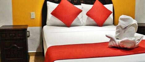 A hotel room with a double bed, red pillows, a red throw blanket, and a towel folded on the bed.
