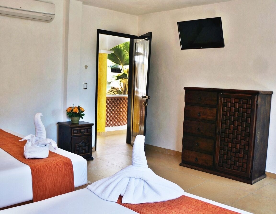 Une chambre d’hôtel comprenant un lit, une armoire en bois, un téléviseur fixé au mur et une porte donnant sur un balcon agrémenté de plantes.