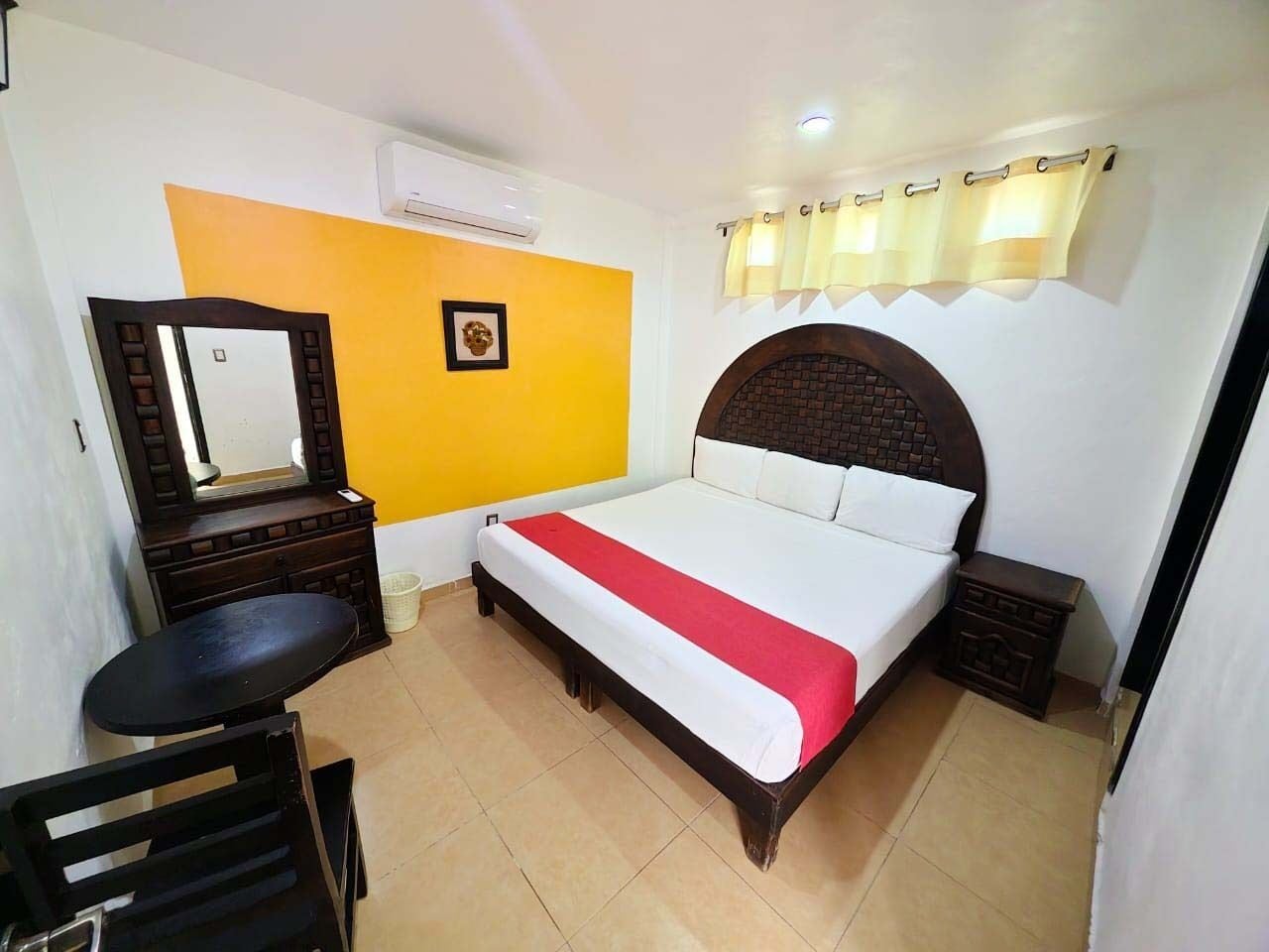 A hotel room with a bed, a small table, a chair, a mirror, and a picture on the wall.