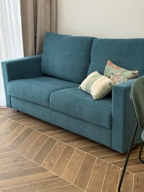 Ein lindgrünes Sofa mit zwei gemusterten Kissen, ein Holzboden mit Fischgrätmuster und ein Sessel mit dunklem Gestell.