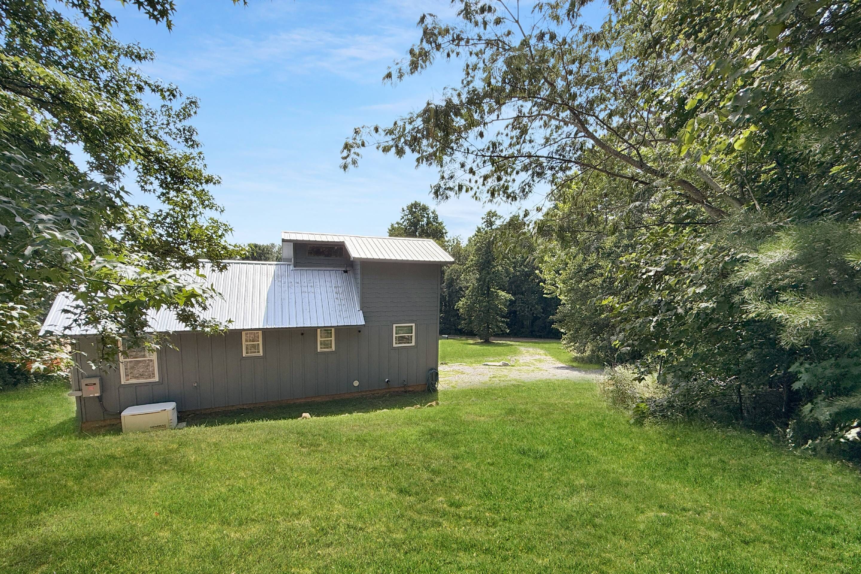 A small, single-story building with a sloped roof, surrounded by a grassy yard and dense foliage.
