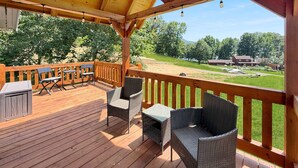 Una terraza de madera con mobiliario exterior, un espacio cubierto y vistas a un paisaje con césped, árboles y un edificio a lo lejos.