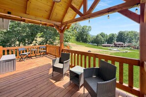 Eine Holzterrasse mit Gartenmöbeln, einem überdachten Bereich und Blick auf eine grüne Landschaft mit Bäumen und einem Gebäude in der Ferne.