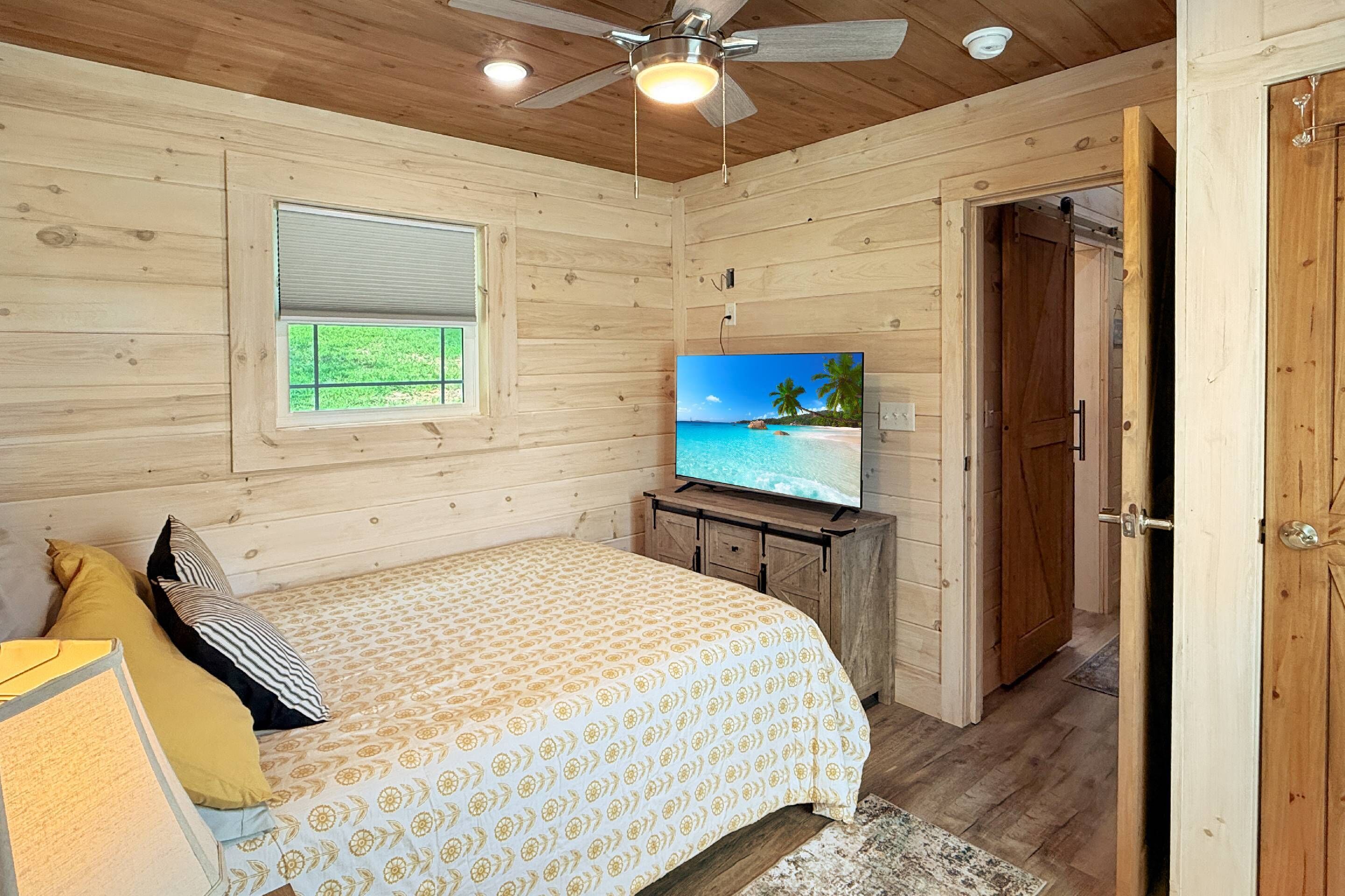 A bedroom with a bed, a television, a ceiling fan, and a window.