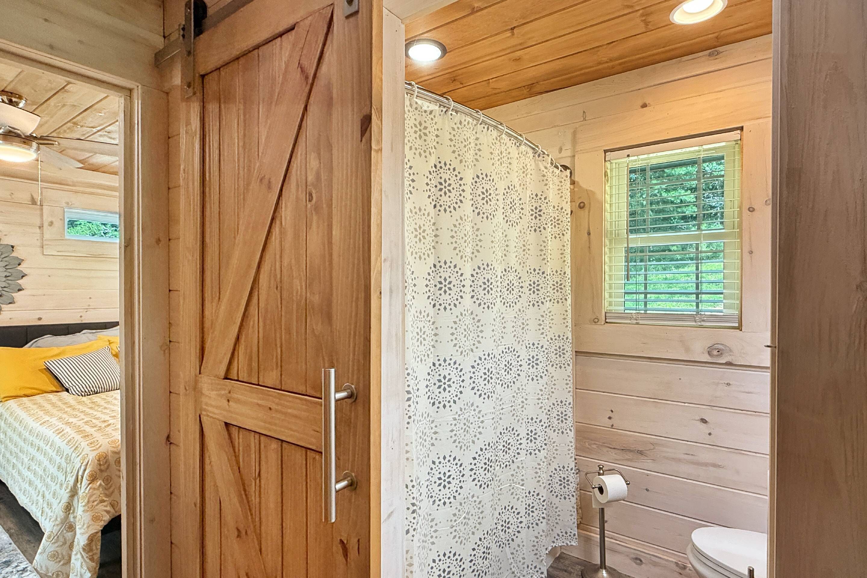 A wooden interior with a bed, a shower with a patterned curtain, and a window with blinds.