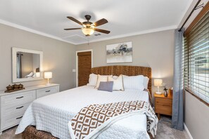 A bedroom with a bed, a ceiling fan, a bedside table with a lamp, a mirror, and a window with blinds. - The River's Edge - Private Home with River Access, Spa, and Karaoke (Three Rivers)