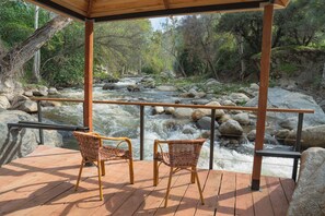 A wooden deck with wicker chairs overlooking a flowing river with rocks. - The River's Edge - Private Home with River Access, Spa, and Karaoke (Three Rivers)