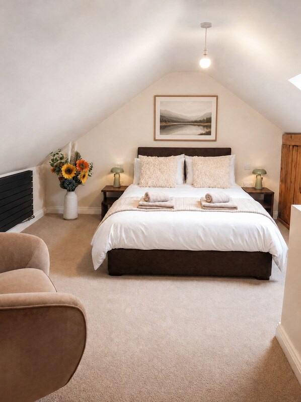 A bedroom with a large bed, two bedside tables, a nightstand, a chair, a wall-mounted lamp, and a framed picture above the bed.