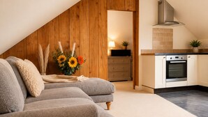 Un moderno living in stile loft con soffitto spiovente, un comodo divano componibile, angolo cottura con fornello e pensili, e un piccolo tavolo da pranzo.