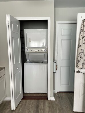 A compact laundry room with a washer and dryer, a door with a floral curtain, and a wooden floor. - Cozy 2-bedroom cottage - St Clair River access in beautiful Algonac with AC (Algonac)