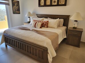 A neatly made bed with a wooden headboard, two bedside tables with lamps, and a view of the outside through a large window. - 360 view of Cerritos Beach, come experience everything Baja has to offer. (El Pescadero)