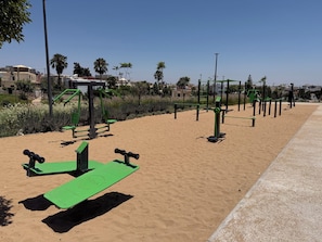 An outdoor exercise area with various fitness equipment, a paved walkway, and a sandy area.