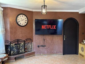 A living room with a wall-mounted clock, a flat-screen TV displaying 'NETFLIX,' a door, and a decorative metal bench. - Charming 3-bedroom house in Agadir Near the Beach perfect for your getaway (AGADIR)