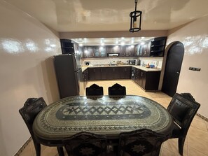 A dining area with a round table and chairs, a kitchen in the background, and a black door. - Charming 3-bedroom house in Agadir Near the Beach perfect for your getaway (AGADIR)