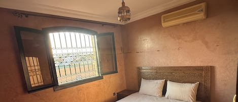 A bedroom with a bed, a window with bars, a ceiling fan, and an air conditioning unit.